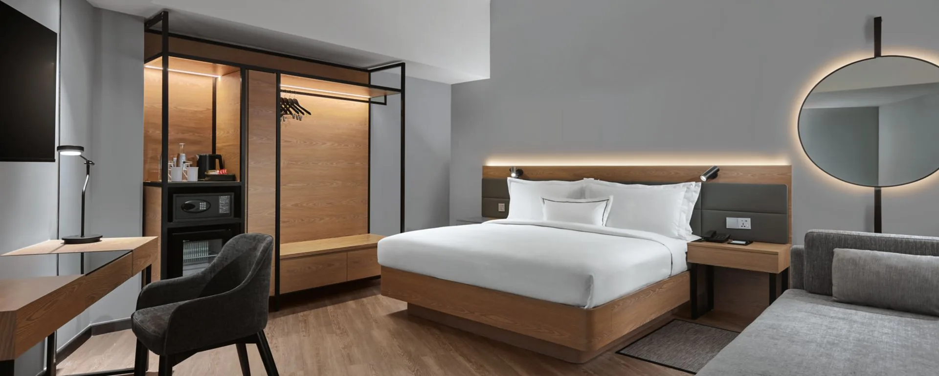 Guest room with king bed, open wardrobe with coffee station and safe, desk with chair, backlit round mirror and sofa